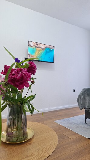 Ett modernt vardagsrum med ett runt träbord, en genomskinlig glasvas med blommor och en platt-TV som är monterad på väggen.