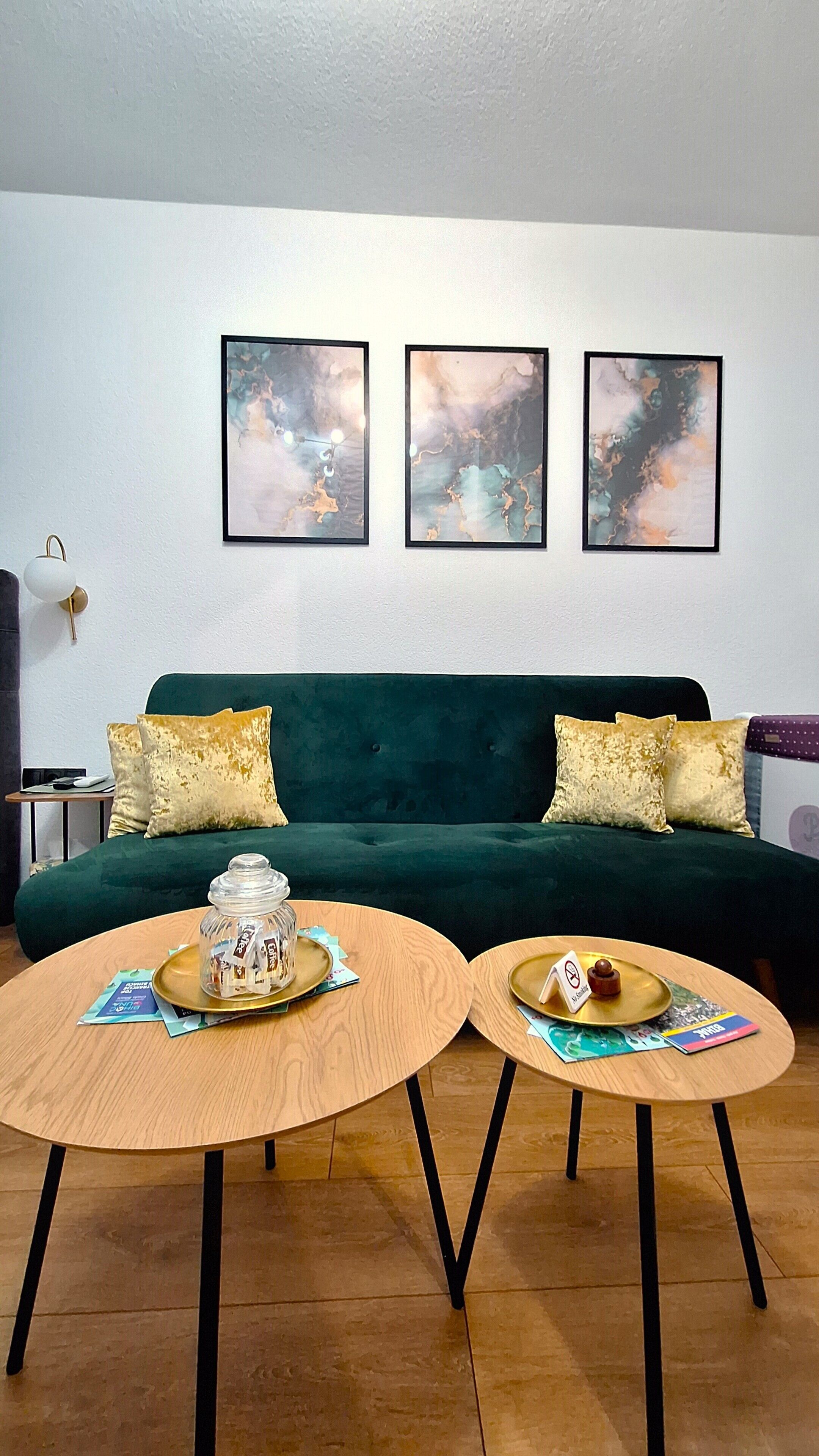 Un salon avec un canapé vert, deux tables rondes en bois et trois tableaux abstraits accrochés au mur.
