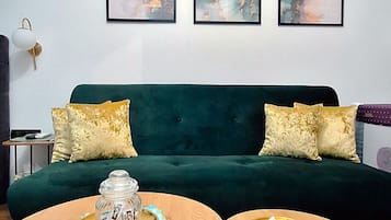Un salon avec un canapé vert, deux tables rondes en bois et trois tableaux abstraits accrochés au mur.