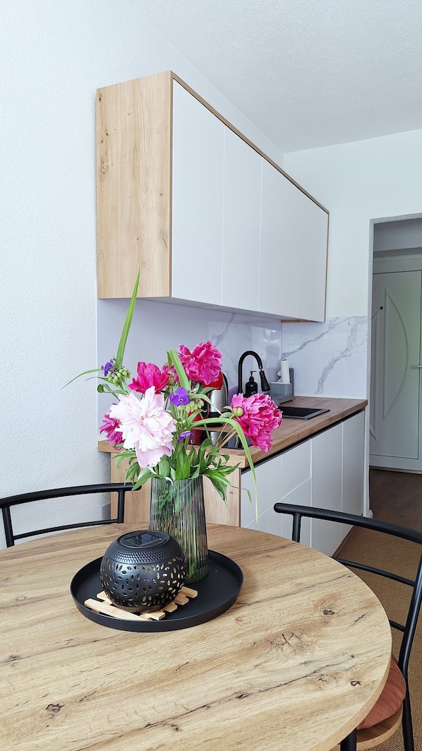 A dining area with a round wooden table, a vase with flowers, a black decorative item, and a kitchen in the background.