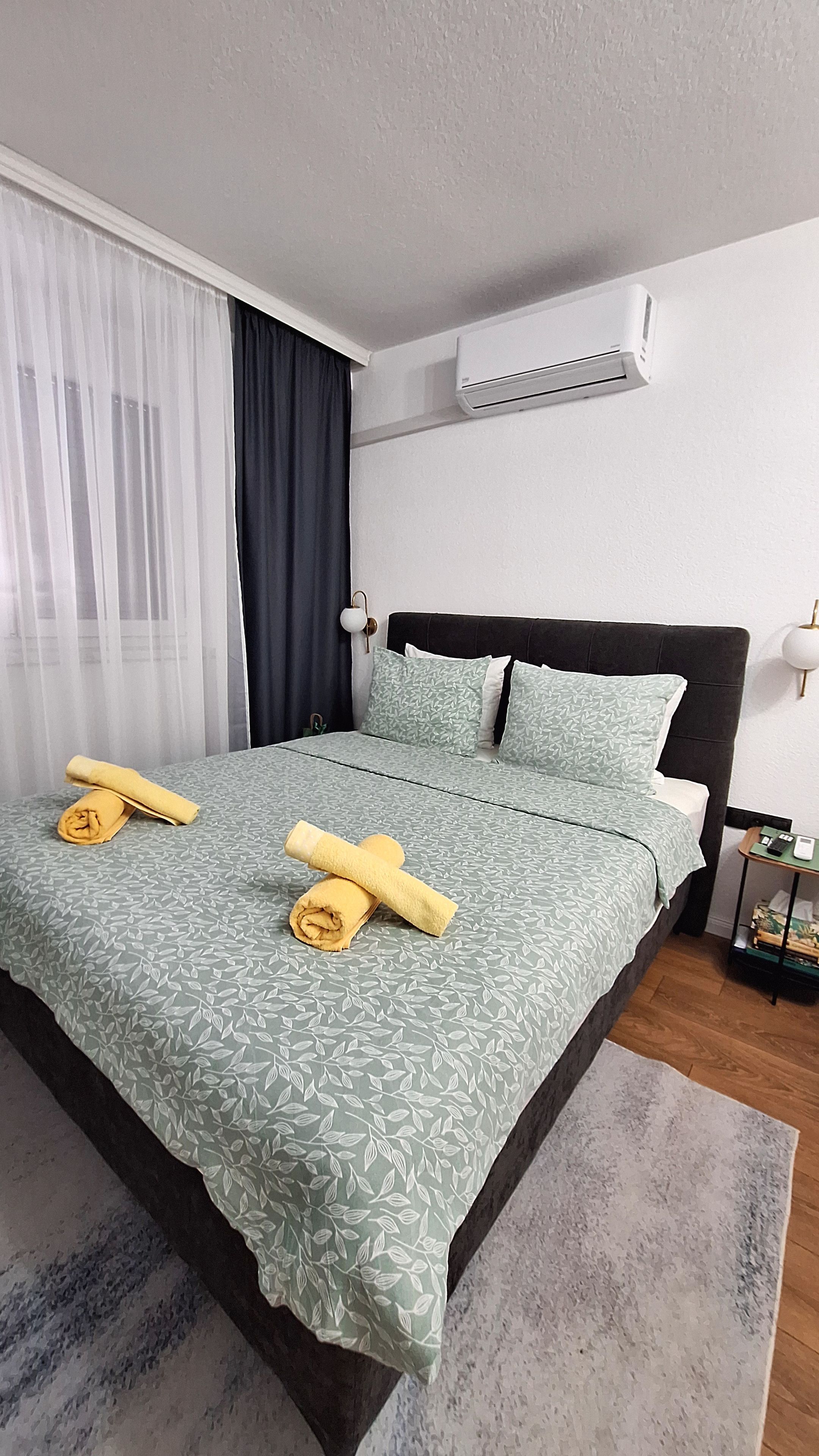 Een opgemaakt bed met een gestreept beddekleed en kussens, een nachtkastje met een lamp, en een airconditioning op de muur.