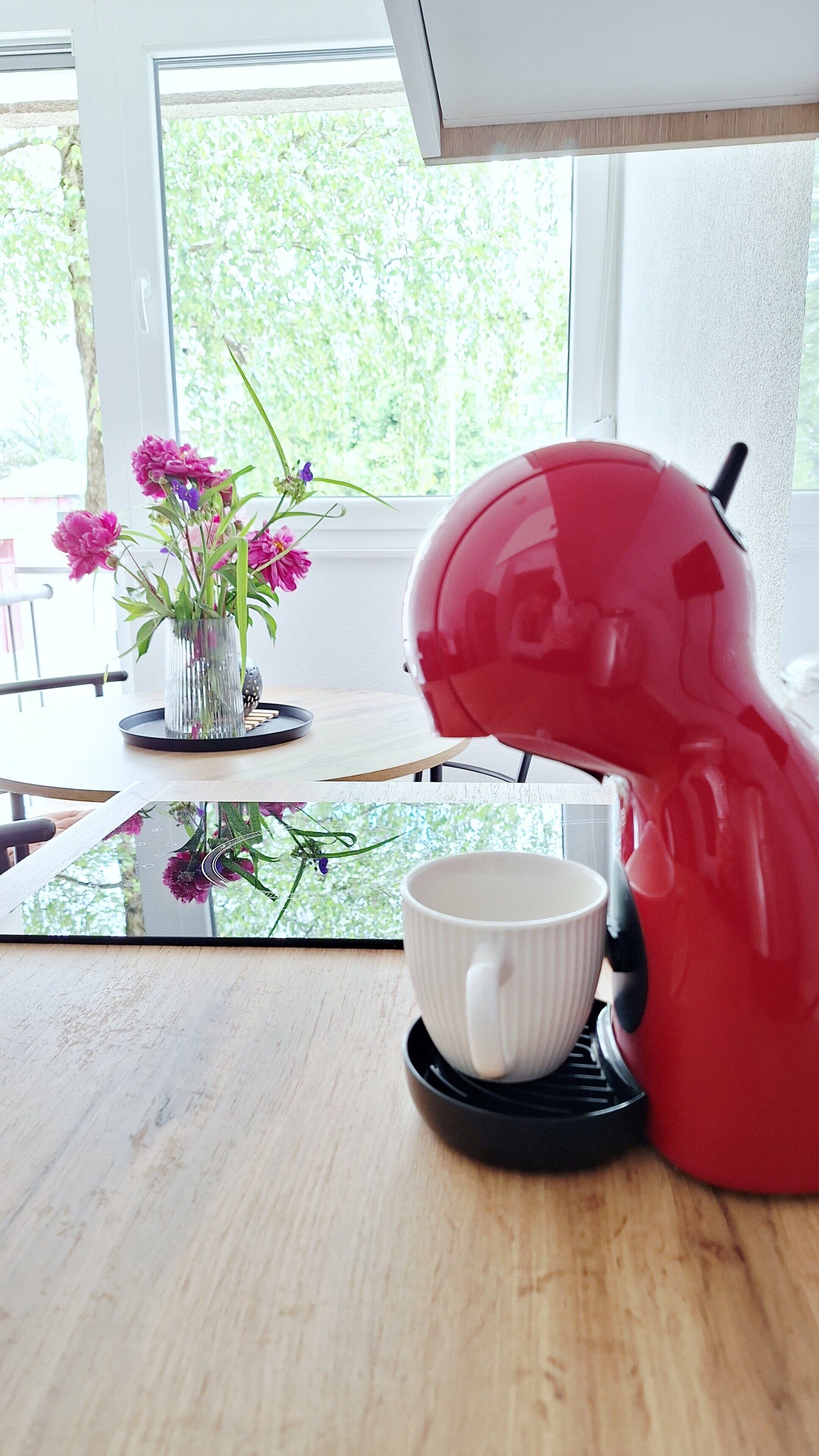 Una cafetera roja sobre una mesa con una taza blanca y un florero con flores.