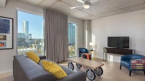 Uma sala de estar moderna com um sofá cinzento, almofadas amarelas, uma mesa de centro de madeira com rodinhas, uma poltrona azul, uma televisão e uma janela grande com vista para a cidade.