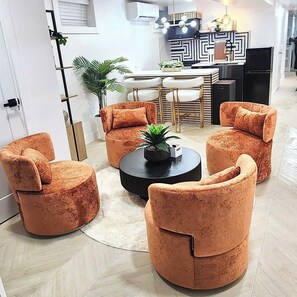 Ein modernes Wohnzimmer mit rundem Couchtisch, vier orangefarbenen Sesseln, einem Esstisch mit Stühlen und einer Küchenzeile mit geometrischer Spritzschutzleiste.