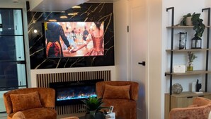 A modern living room with a large flat-screen TV, two brown velvet armchairs, a black coffee table, and a fireplace. - Luxe 3BR with 3 Private Bathrooms Near Airport and 30 mins to NYC (Newark)