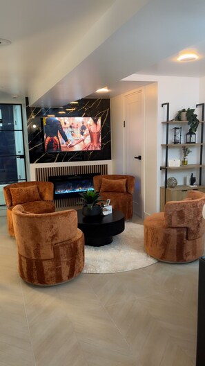 A modern living room with a large flat-screen TV, two brown velvet armchairs, a black coffee table, and a fireplace. - Luxe 3BR w/Private Bath in Every BR Near Airport (Newark)