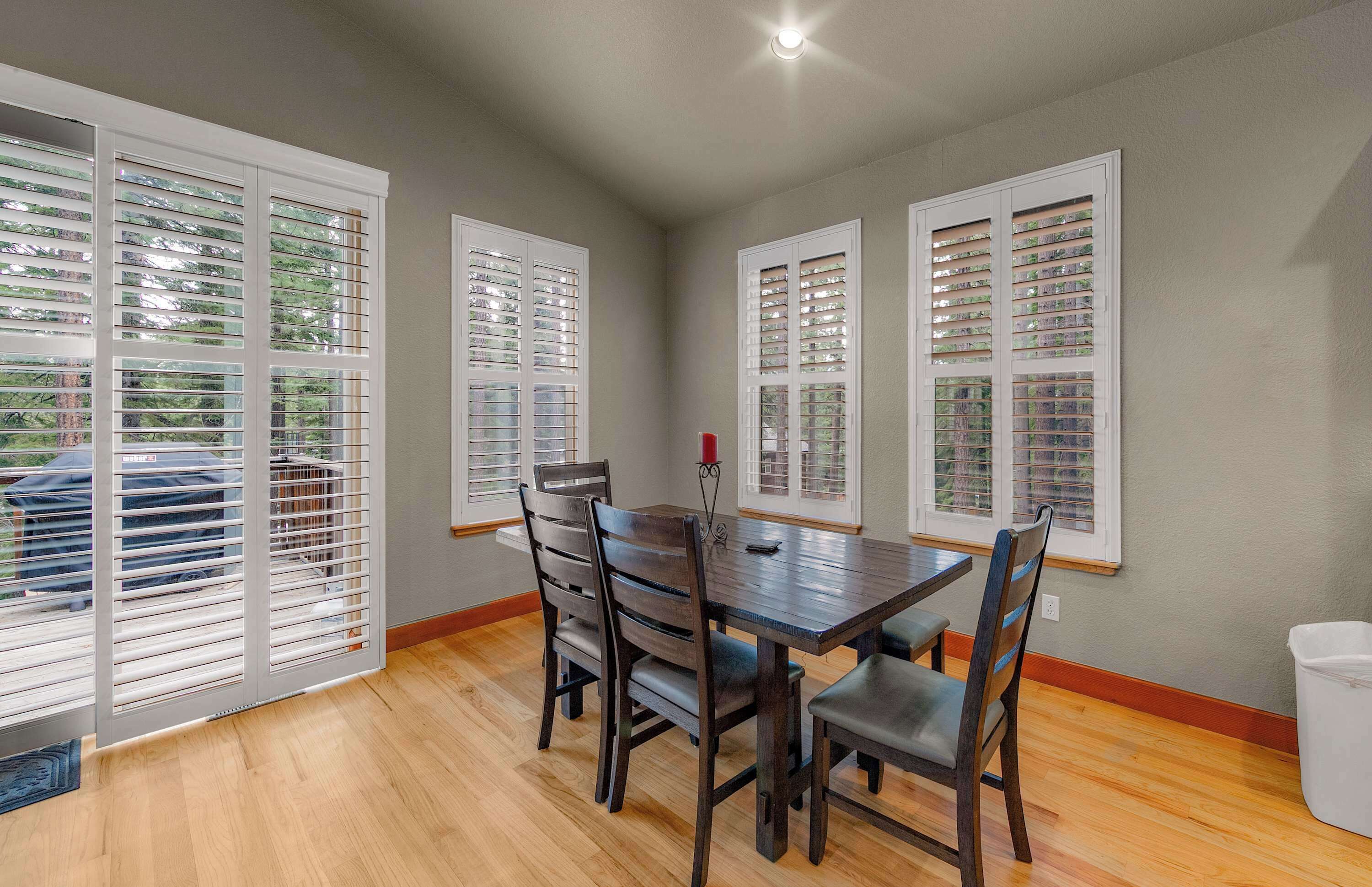 A dining area with a wooden table and chairs, large windows with blinds, and a ceiling light.