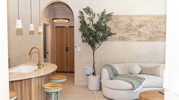 Ein modernes Badezimmer mit Waschbecken, Ablagefläche und Hocker. Im Wohnbereich befindet sich eine Pflanze und ein Sofa.