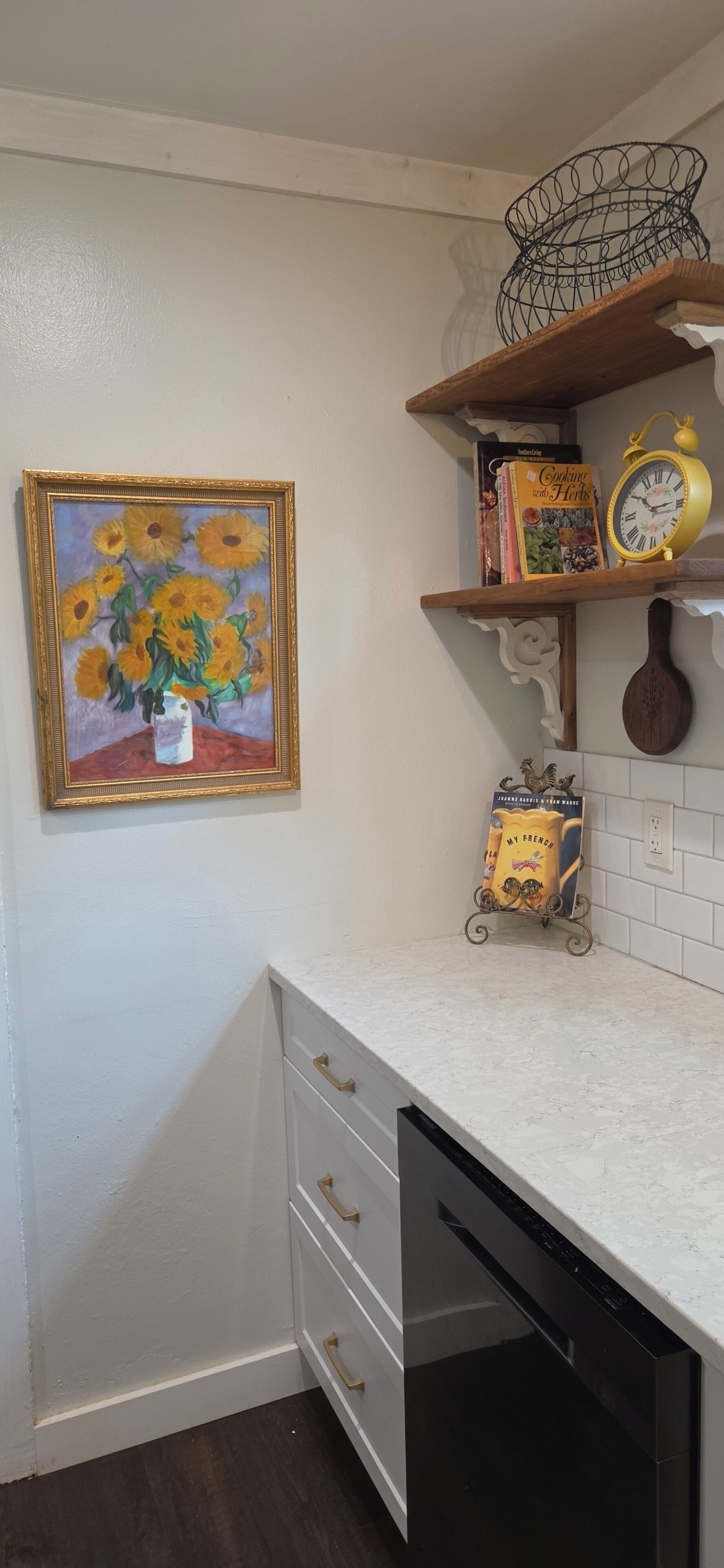 Una cocina con encimera blanca, lavavajillas, un cuadro enmarcado de girasoles y una estantería con libros y un reloj.