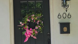 Een zwarte deur met een roze bloemenkrans, een brievenbus met het nummer 606, en een deurmat.