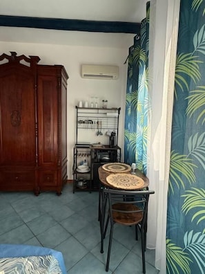 A room with a wooden cabinet, a small table with chairs, a metal rack with kitchen items, and a patterned curtain.