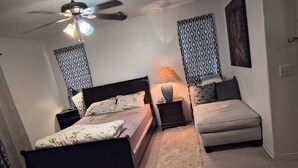 Une chambre à coucher comprenant un lit, une table de chevet, une lampe, un canapé, un ventilateur de plafond et des rideaux à motifs.