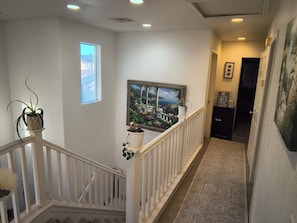 A hallway with a white railing, a potted plant, a framed picture, and a door leading to another room.