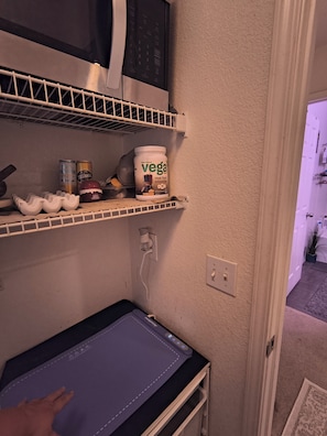 A kitchen with a microwave, a shelf with a plant-based smoothie container, a coffee maker, and a baby changing station.