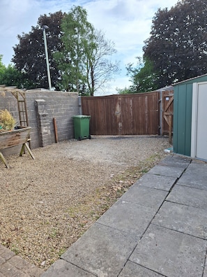 A backyard with a gravel area, a wooden fence, a green trash bin, and a shed.