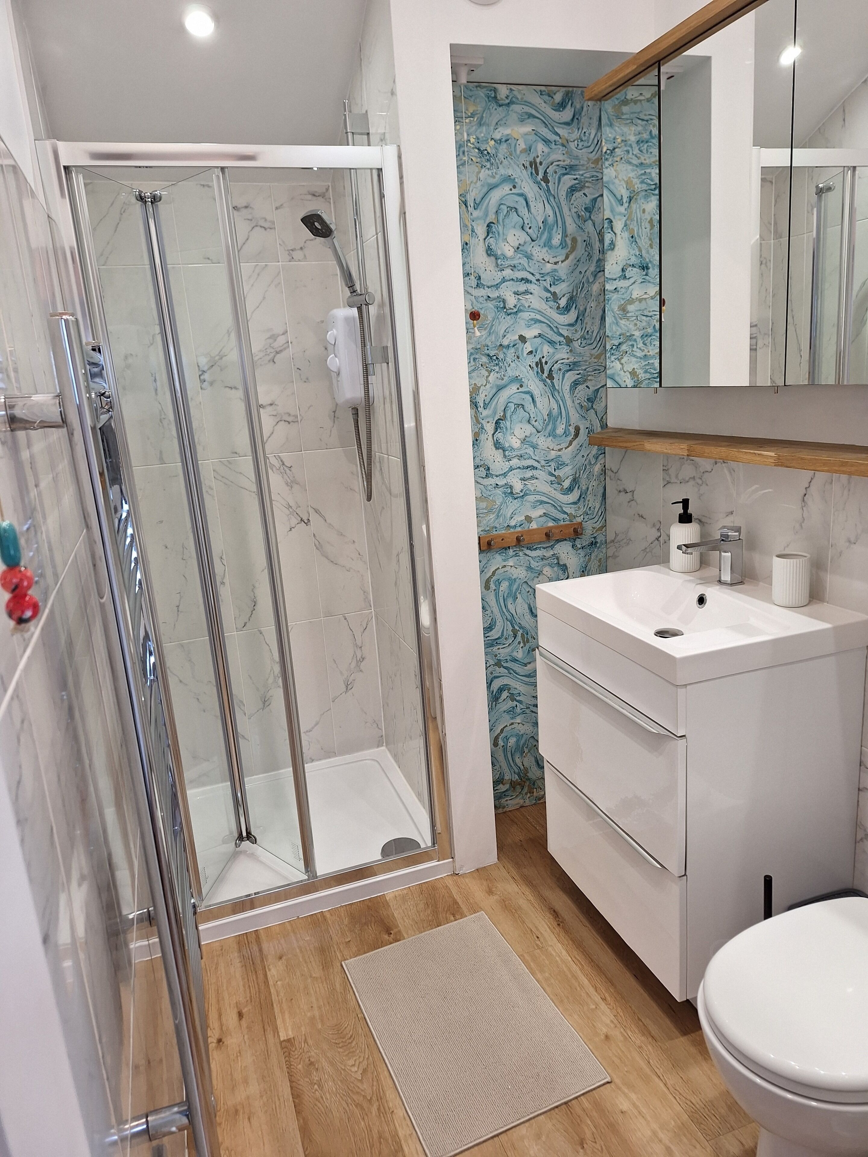A bathroom with a glass-enclosed shower, a marble-tiled floor, a wooden floor mat, a white sink with a mirror above, and a marble-patterned wall.. Cosy Garden Cottage, Combe Down | Free Parking