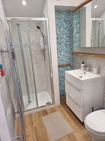 A bathroom with a glass-enclosed shower, a marble-tiled floor, a wooden floor mat, a white sink with a mirror above, and a marble-patterned wall.. Cosy Garden Cottage, Combe Down | Free Parking