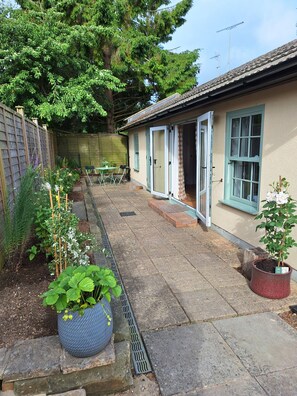 A residential property with a paved patio, a small garden with various plants, a wooden fence, and a building with a tiled roof and a door. - Cosy Garden Cottage, Combe Down | Free Parking (Bath and North East Somerset)
