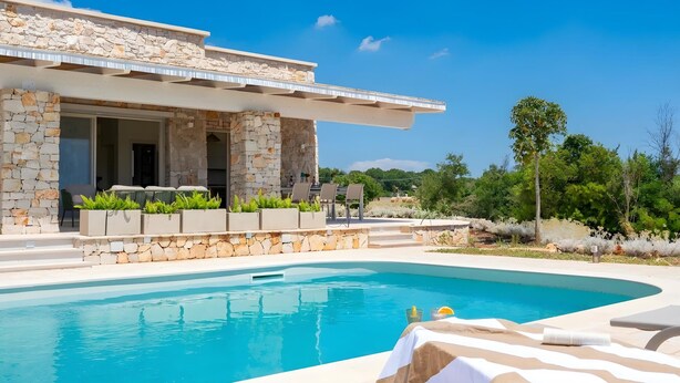 A modern villa with a stone facade, a swimming pool, outdoor seating, and a view of the surrounding landscape. - High-end pool villa on 2 levels with sea view (Torre Pali)
