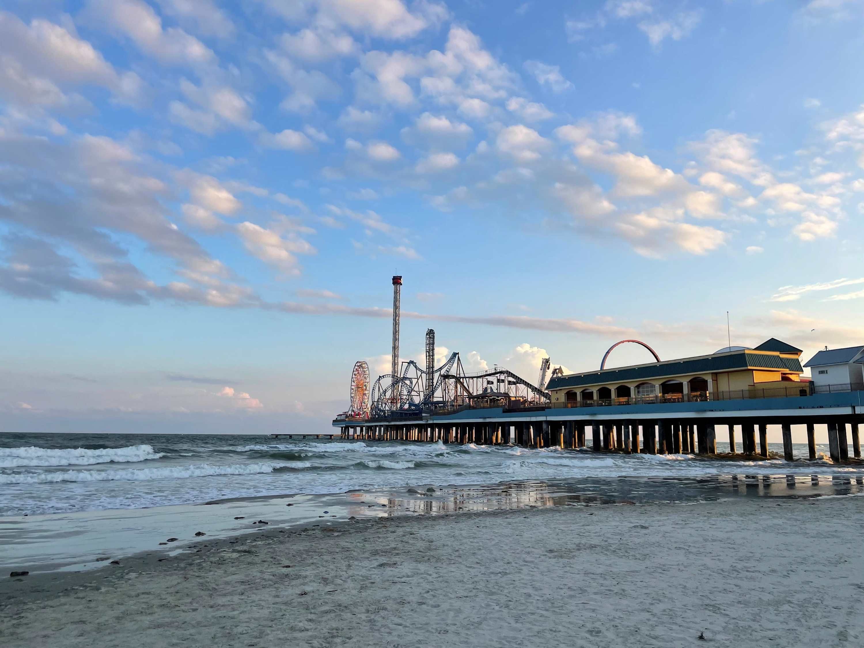 A beach with a pier featuring amusement rides, a building with a flat roof, and the ocean in the background.