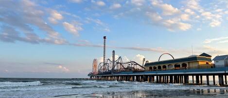 A beach with a pier featuring amusement rides, a building with a flat roof, and the ocean in the background.