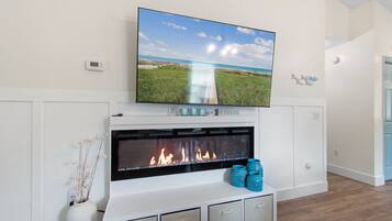 A modern living room with a fireplace, a flat-screen TV mounted above it, and a view of a beach through the TV screen.