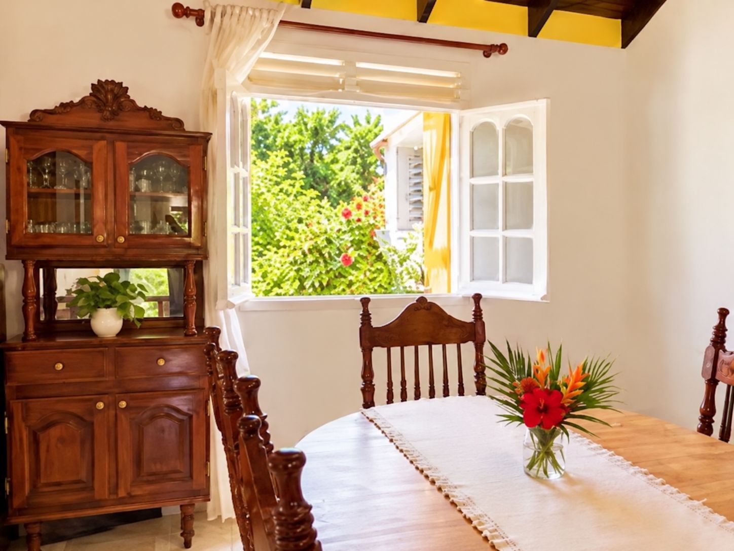 A room with a dining table, a wooden cabinet with glass doors, and a window with an open view of greenery.