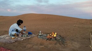 Una persona sentada sobre una manta en un paisaje desértico, con fuego y una botella de agua.