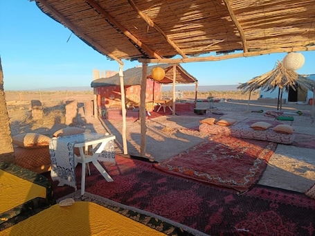 A rustic outdoor seating area with a thatched roof, a small table, and colorful rugs.. Desert Erg Chigaga