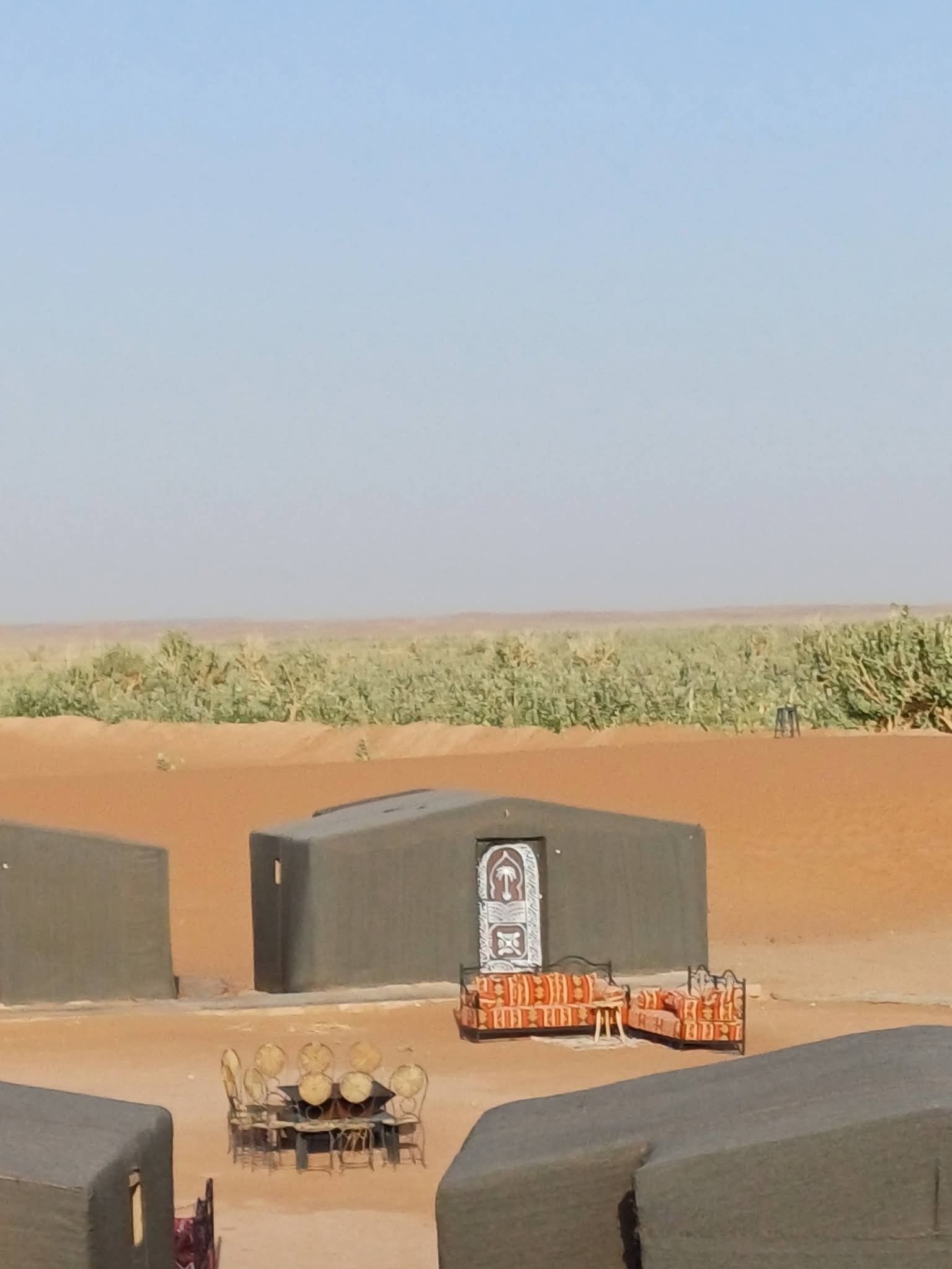 Un paysage désertique, avec un ciel dégagé, une rangée de tentes et, au premier plan, une table entourée de chaises.
