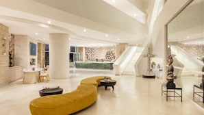 Eine großzügige Lobby mit einer zentralen Rezeption, einer gebogenen gelben Couch und einer Treppe zu den oberen Stockwerken.