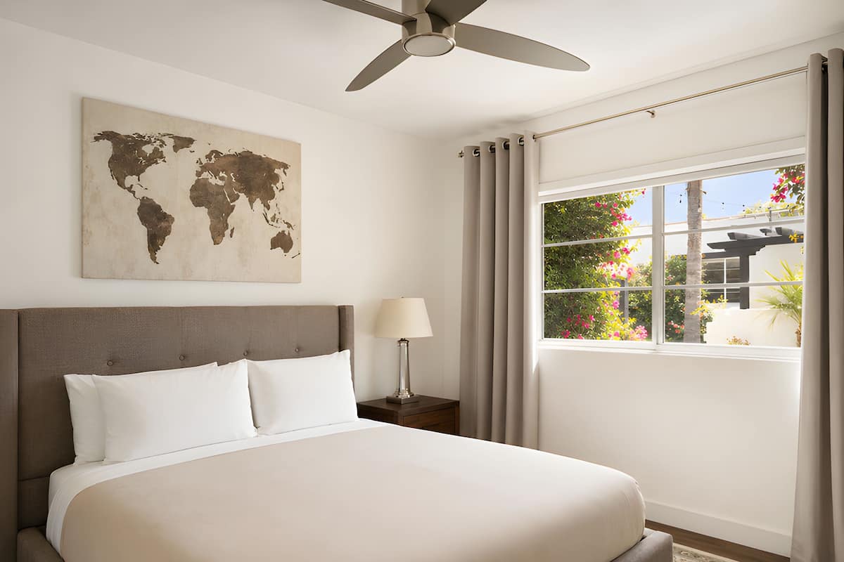 A hotel room with a large bed, a world map on the wall, a ceiling fan, a window with curtains, and a nightstand with a lamp.