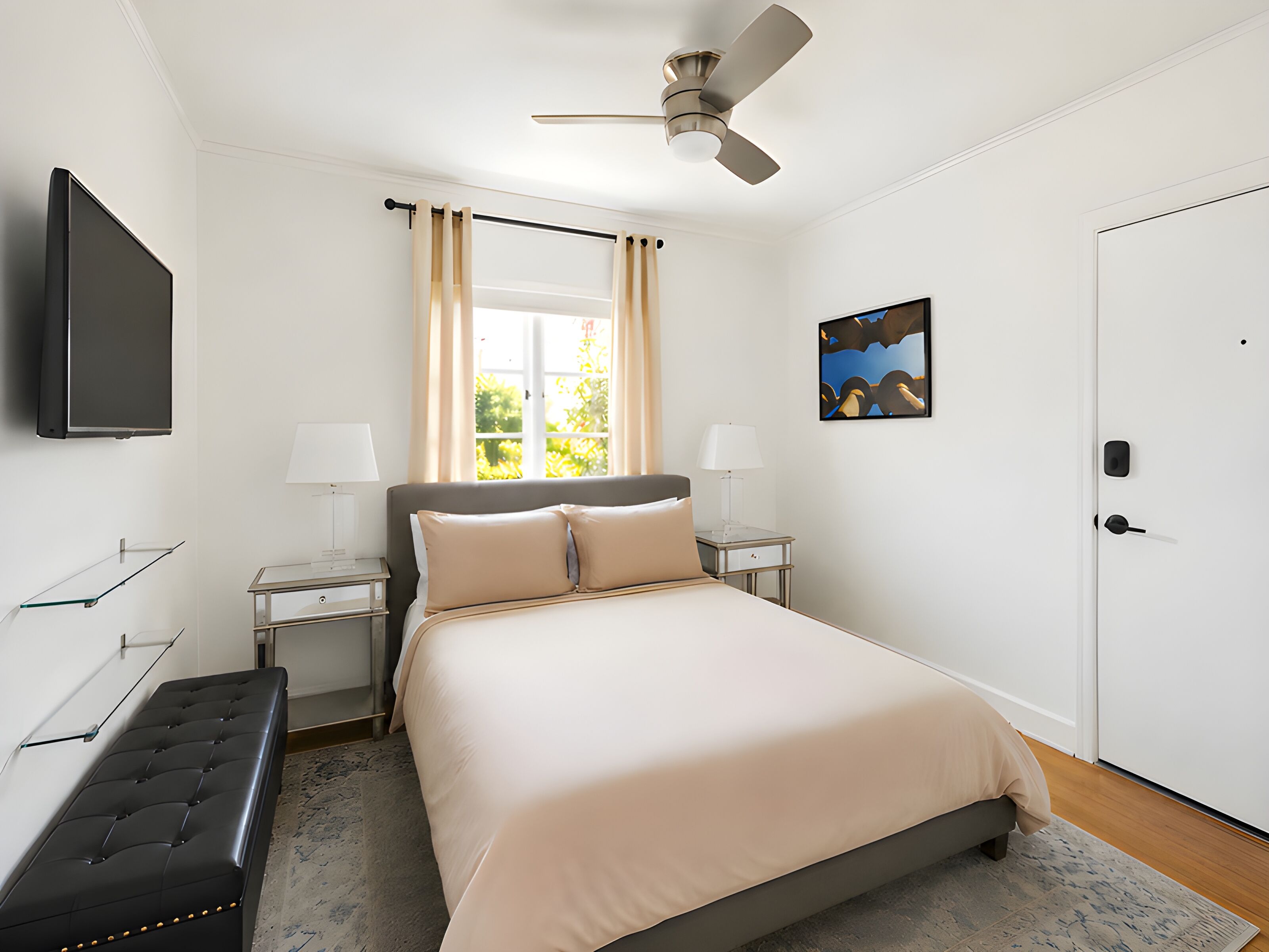 Un dormitorio con cama, dos mesitas de noche, ventilador de techo, televisor, banco, ventana con cortinas y puerta.