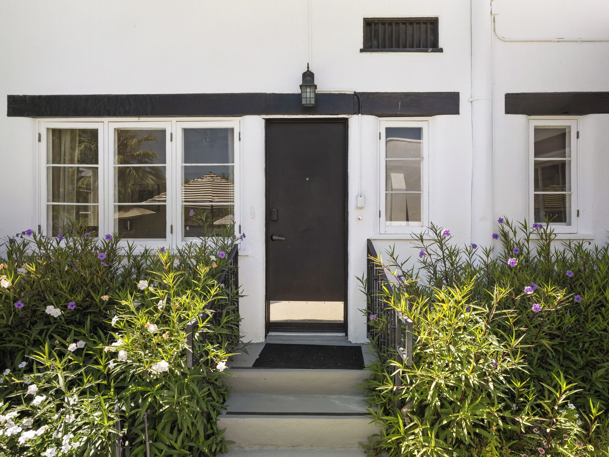 A white building with a black door, windows, and a small lamp above the door. There is a pathway leading to the door, flanked by plants with purple flowers.