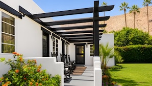 A covered patio with outdoor seating, a white building, and a well-maintained garden with palm trees and flowering plants. - Gloria Palm Springs (Palm Springs)