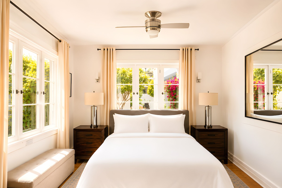 A hotel room with a large bed, two bedside tables, a ceiling fan, and large windows with curtains.