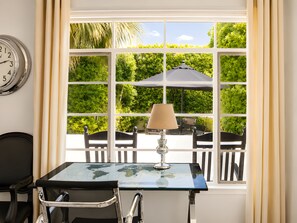 Una habitación con una mesa, sillas, una lámpara, un reloj y una ventana que da a zonas verdes.