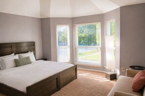 A bedroom with a large bed, two windows with blinds, a small table, and a chair. - Plano Luxury Retreat · Hot Tub · Theater · Game Room Dallas/Frisco Sleeps 8 (Plano)