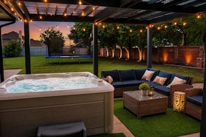 Een hottub, een buitenbank en rieten meubels onder een pergola met lichtslingers.