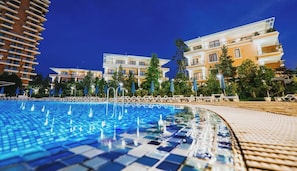 A swimming pool with a tiled edge, surrounded by lounge chairs and umbrellas, with buildings in the background.