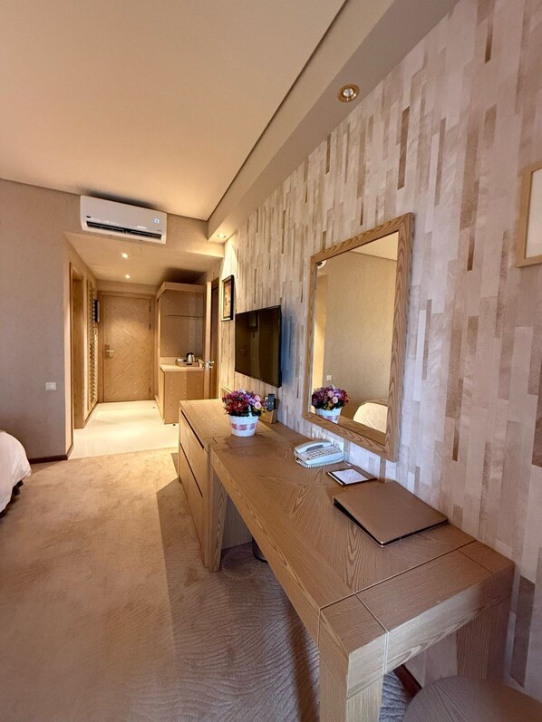 Habitación de hotel con un escritorio de madera, un espejo grande, un televisor de pantalla plana y un baño pequeño con ducha.