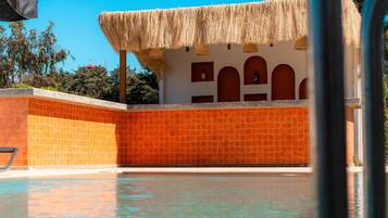 Un área de piscina con un muro de azulejos, una estructura de techo de paja y un cielo azul despejado.