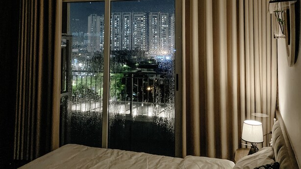 A hotel room with a bed, a nightstand, a lamp, and a view of a cityscape through a large window. - Compact Studio w/ Projector & Sky View (Hà Nội)