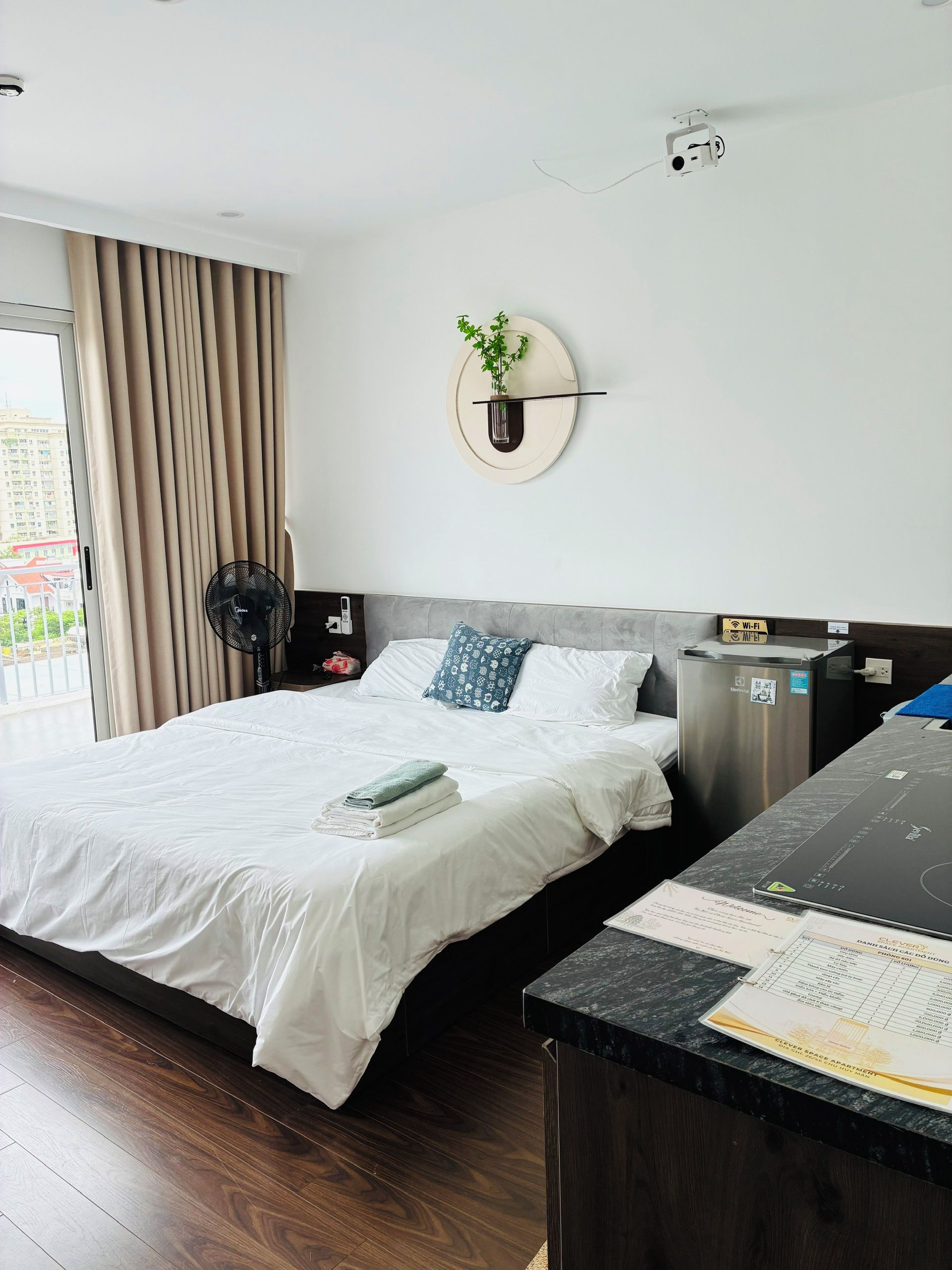 A hotel room with a bed, a desk, a plant, and a view of the outside.