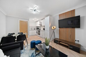A modern living room with a dining area, a kitchen, and a TV mounted on a wooden panel wall. - Spacious 3-Bed Apartment • Sleeps 6 • Free Parking • 15mins to Central London (London)