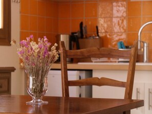A dining table with a vase of dried flowers, a wooden chair, and a kitchen area with orange tiles in the background. - Les Lilas Apartment (2–4 people) (BASSURELS)
