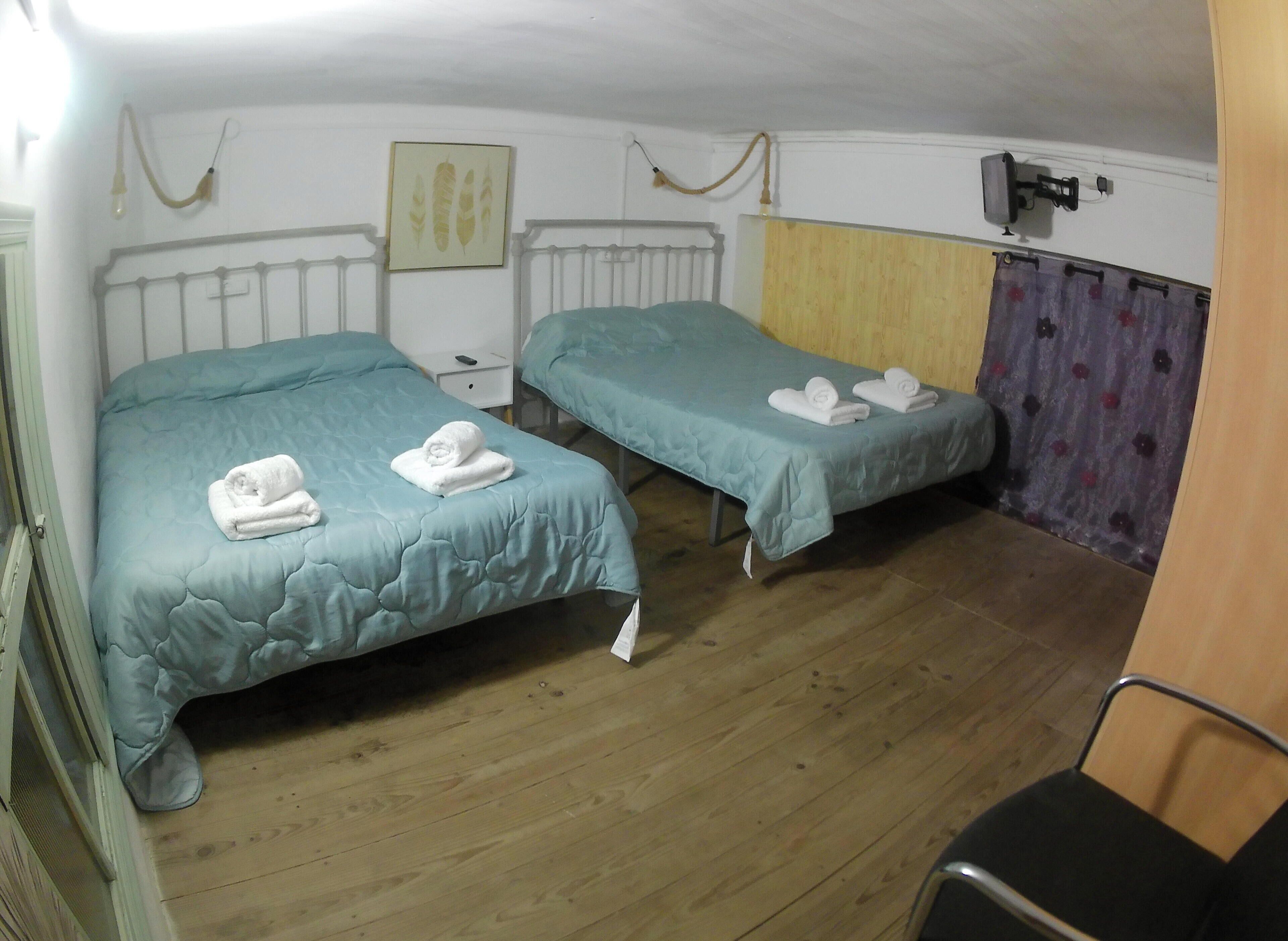 Habitación con dos camas, cada una con una colcha verde azulada y toallas blancas. El suelo es de madera, hay un radiador blanco y un televisor montado en la pared.
