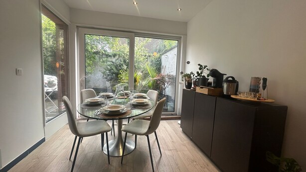 A modern dining area with a glass table, white chairs, and a view through large windows.