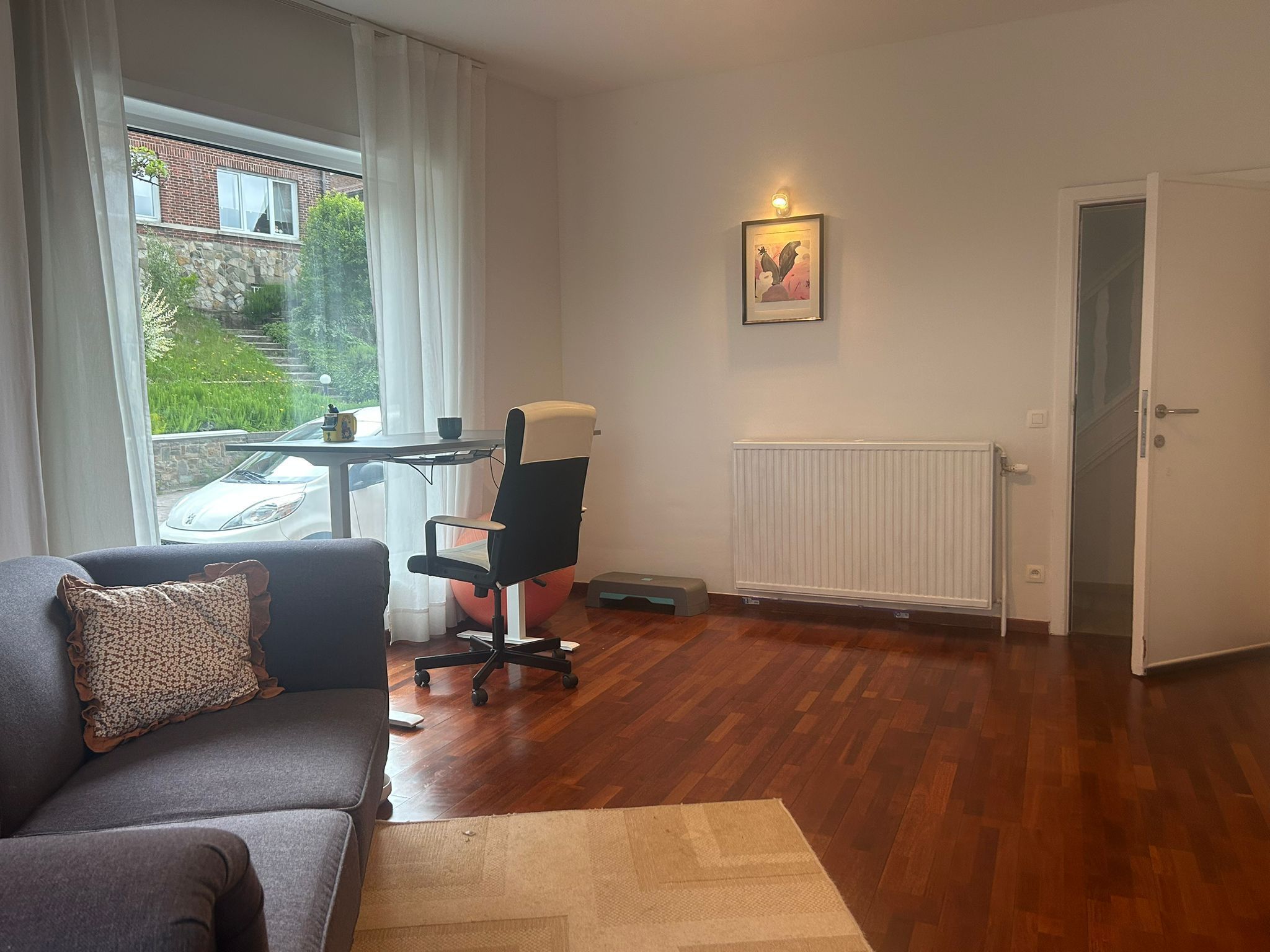 A living room with a sofa, a chair, a table, a radiator, and a window with a view of a building and greenery.