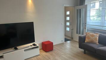 A modern living room with a flat-screen TV, a red storage box, a grey sofa with a patterned cushion, and a door leading to another room.